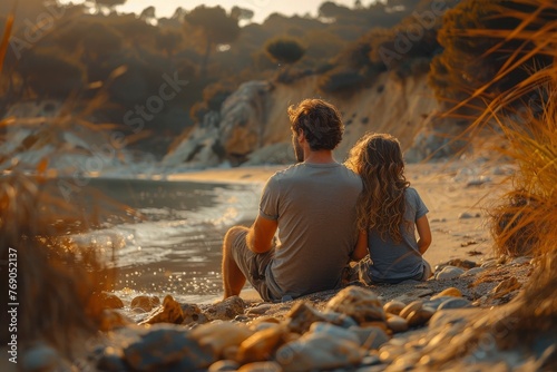 A couple sits closely together  looking at a tranquil sea against a backdrop of a beautiful sunset and rocky beach