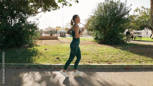 beautiful fair-haired European woman goes for a run in a green city park on an early summer morning, shaded by trees.camera moves alongside her, capturing scene in slow motion.
