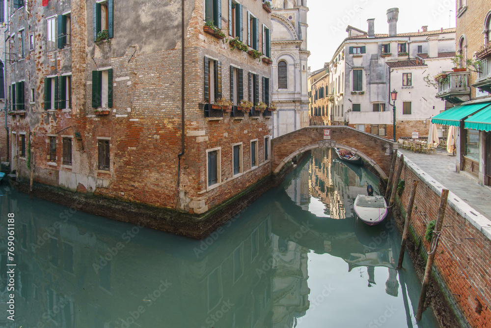 Typical narrow canal surrounded by buildings with boats and a footpath bridge in Venice, Veneto, Italy