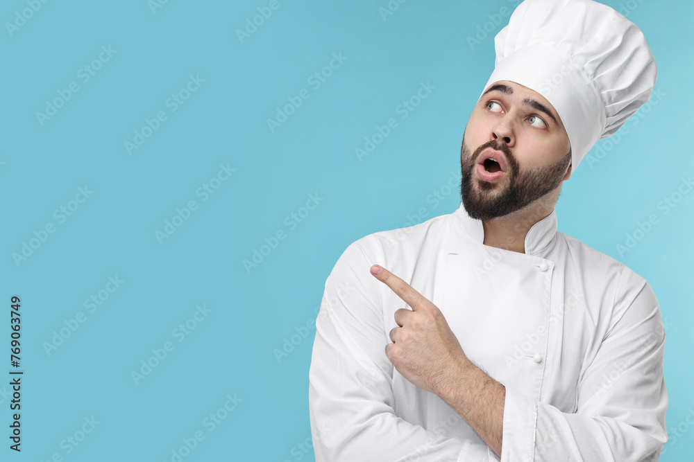 Surprised young chef in uniform pointing at something on light blue background. Space for text Surprised young chef in uniform pointing at something on light blue background. Space for text