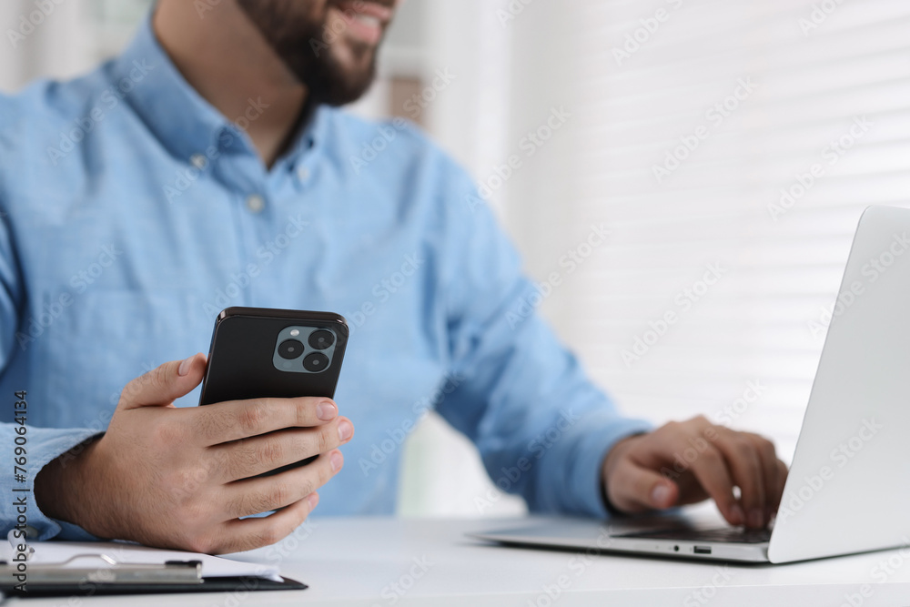 Young man using smartphone while working with laptop at white table in office, closeup