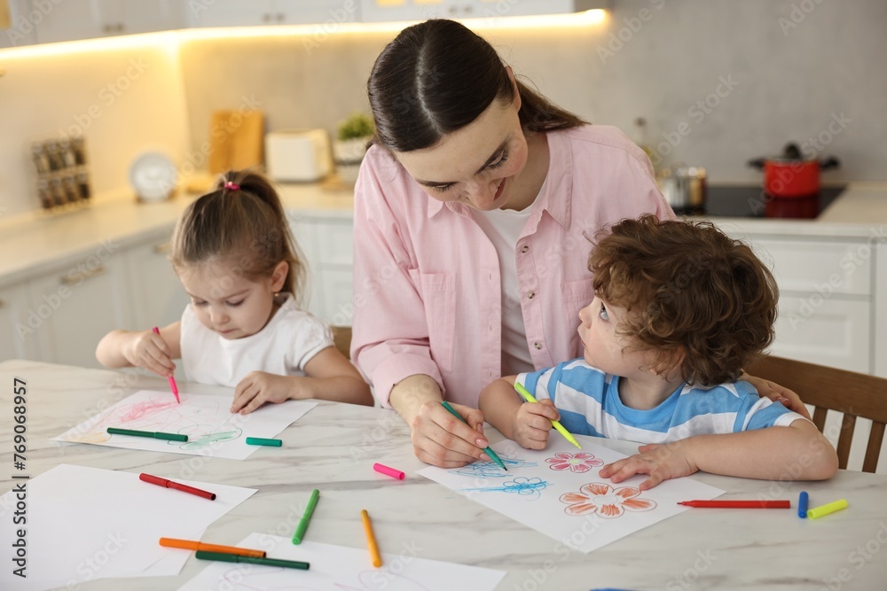 Mother and her little children drawing with colorful markers at table in kitchen