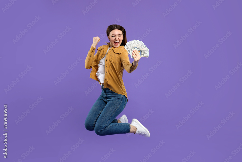 Excited woman with dollar banknotes jumping on purple background