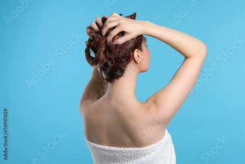Young woman washing her hair with shampoo on light blue background, back view
