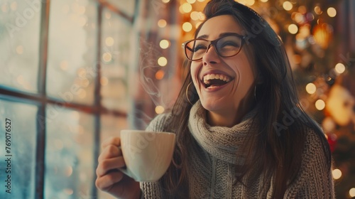 Portrait of joyful young woman enjoying a cup of coffee at home. Smiling pretty girl drinking hot tea in winter. Excited woman wearing spectacles and sweater and laughing in an autumn day
