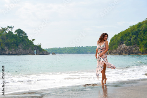 A slender young woman in a pink light dress walks along a tropical beach. The girl is dancing in the sea waves.