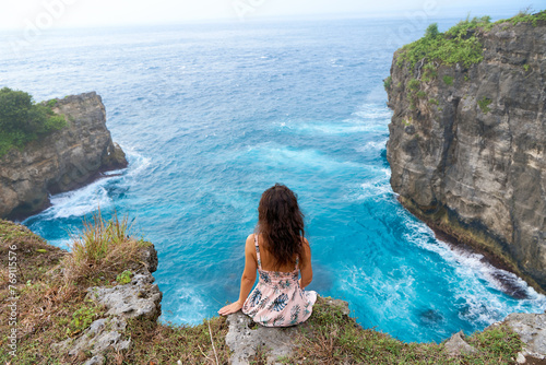 A beautiful woman in a pink dress sits on a cliff above the ocean on the island of Nusa Penida. Devil's Billabong an incredibly wonderful lagoon with splashes from the waves.