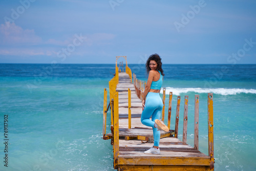A slender young woman walks along an old wooden pier, which goes into the horizon of the incredible azure color of the ocean.