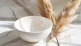   A white bowl rests on a white counter, adjacent to a tall, dried grass plant