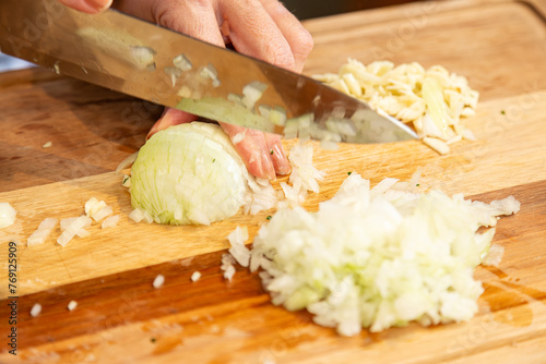 Chef com faca picotando e preparando cebola e alho na tabua de corte na cozinha.	
 photo