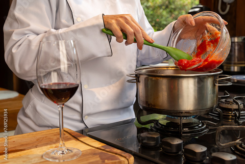 Chef colocando molho de tomate na panela enquanto toma um vinho tinto, preparando uma receita italiana a beira do fogão	
 photo