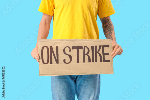Protesting young man holding placard with text ON STRIKE against blue background, closeup © Pixel-Shot
