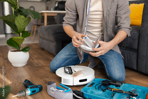a man, a craftsman, repairs and routinely inspects a robot vacuum cleaner after operation on a wooden floor with work tools. A cozy room with a gray sofa and green flower pots. cleaning the air filter photo
