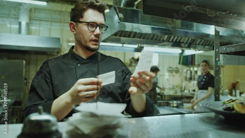Chef taking order tickets from metal rack, reading them and telling what to cook to kitchen crew during workday in restaurant photo