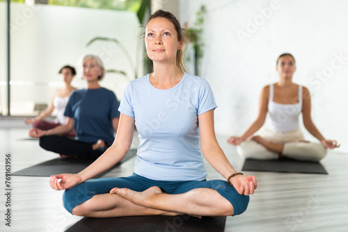 Relaxed middle-aged woman meditating in Lotus Pose Padmasana on mat during group yoga class in fitness studio