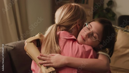 Back slowmo of little girl in glasses and with bionic plastic arm hugging her mother while spending time together at home photo