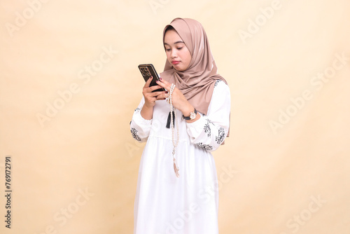 a young indonesia Muslim female wearing a cute hijab is holding prayer beads and WhatsApp via smartphone gadget. for advertising, lifestyle, banners and Ramadan