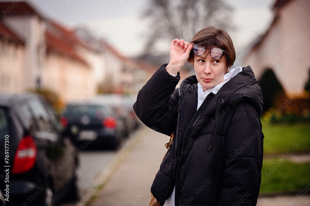 A young woman with a curious expression lifts her glasses on a suburban street, with blurred residential homes in the background