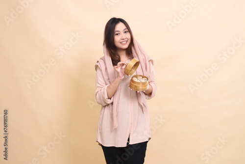 Asian girl in semi-hijab stands happily with her hands opening the lid of a wooden bowl containing dim sum. beautiful Muslim women for culinary, business, food