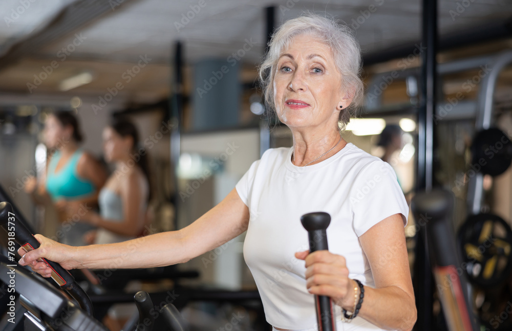 Fototapeta premium Slender athletic mature woman during cardio exercises on elliptical simulator in fitness center. Sports to max. Electronic tracking for most productive workout