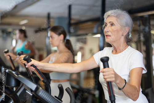 Active athlete senior woman engaged in intensive high interval training on ellipsoid simulator. Training of respiratory and cardiovascular system, increasing endurance in crossfit gym photo