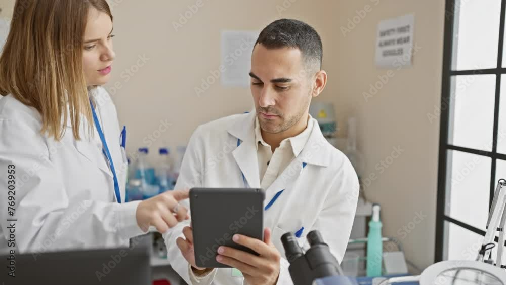 A man and woman in lab coats analyze data on a tablet in a modern laboratory setting. A man and woman in lab coats analyze data on a tablet in a modern laboratory setting.