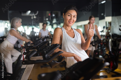 Fototapeta Naklejka Na Ścianę i Meble -  Smiling active young adult woman posing near stationary bike after cardio workout in gym, showing thumbs up gesture