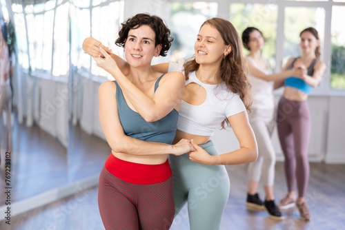 Two smiling girls in activewear performing kizomba in pair, demonstrating dance characteristic sensual and connected movements in studio class setting