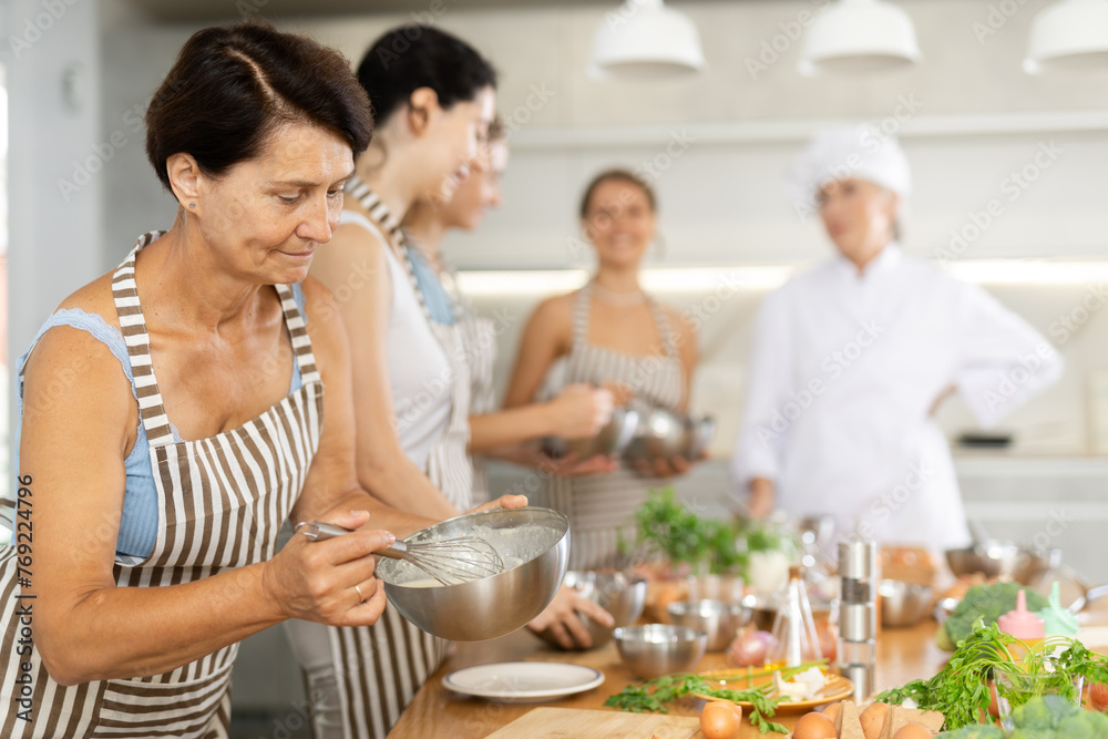 Mature woman at group cooking class holds bowl in hands and mixture cream sauce with white wine for meat with whisk
