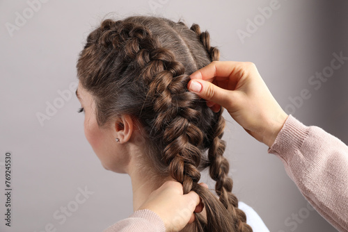 Professional stylist braiding woman's hair on grey background, closeup photo