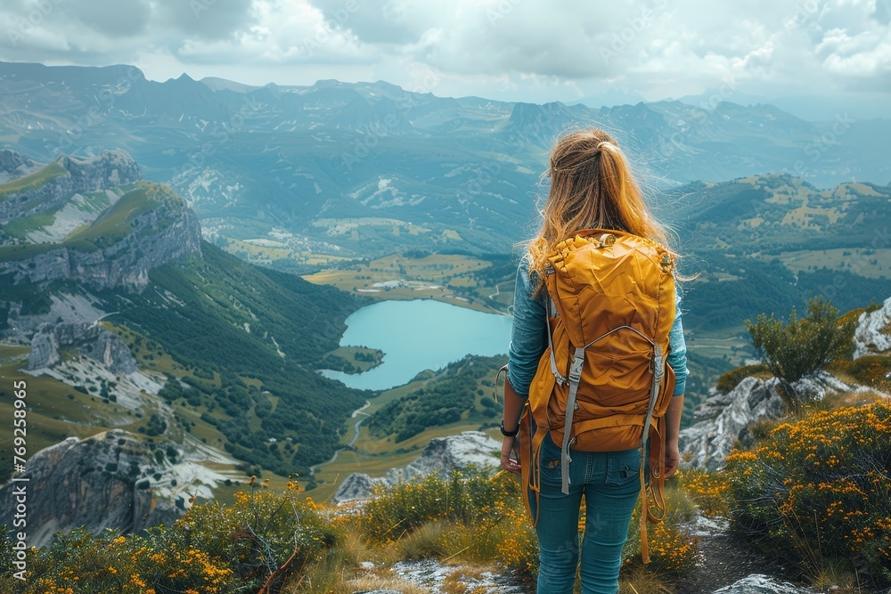 A young woman in a yellow backpack gazes at a distant mountain lake surrounded by rugged peaks and wildflowers