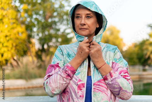 Beautiful senior woman 50-60 years old, wearing colorful clothes, 80s style hoodie, posing, smiling and doing exercises, stretching in an outdoor park at sunset photo