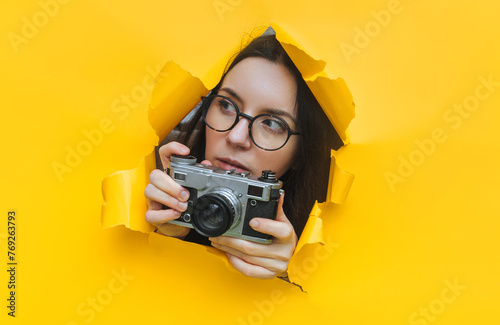 A young paparazzi girl in glasses with a rare SLR camera looks out from her hiding place and carefully watches what is happening. Yellow paper hole.Tabloid press.Looking for a subject for stock photos