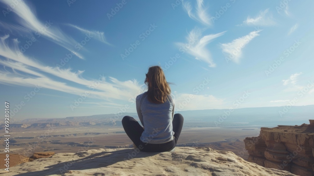 A woman sits crosslegged on a rock back to the camera as gazes towards the endless blue sky and endless stretch of the desert . .