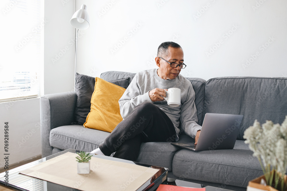 Senior Asian man sitting on sofa and using laptop while holding a cup of coffee in a modern living room Senior Asian man sitting on sofa and using laptop while holding a cup of coffee in a modern living room