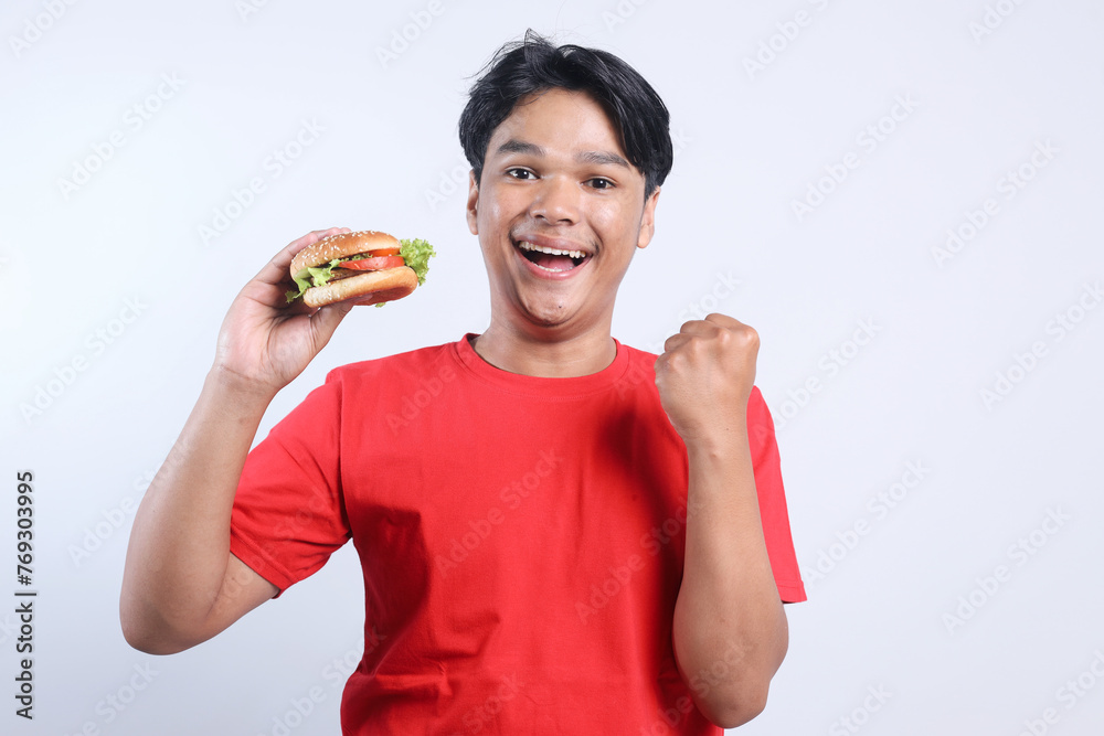Excited Asian man clenching fist while holding hamburger over white background