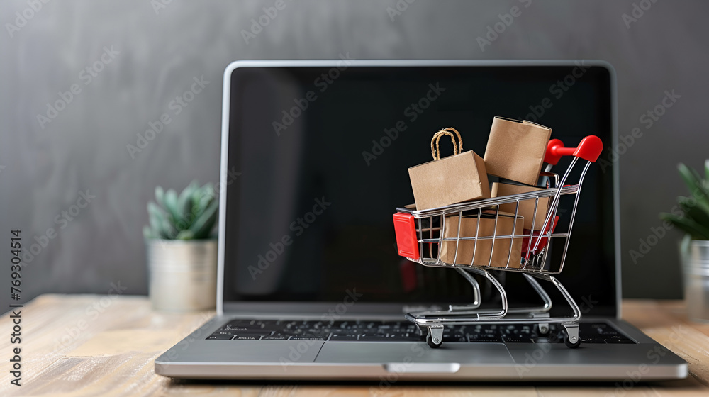 The laptop screen shows a shopping cart full of boxes. Shopping cart on a wooden table and boxes stacked. Online shopping and parcel service concept