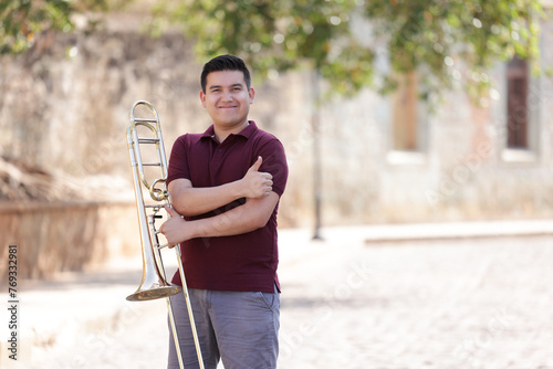 portrait of young mexican man smiling with thumbs up and holding trombone with blurred background photo