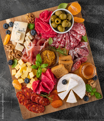 Charcuterie board with a variety of cheeses, salami, chorizzo, prosciutto, honey, grapes, nuts, olives, bread, blueberries and fresh herbs on a dark concrete background. A festive snack.