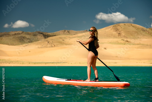 Sporty woman in black swimsuit is standing on stand up paddle sup board at desert lake with turquoise water against sandy dunes