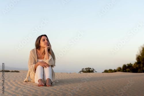 A woman sits peacefully on a sandy dune, her gaze thoughtfully directed towards the horizon as the sunset casts a warm glow on her and the surrounding sand. The tranquil setting suggests moments of in