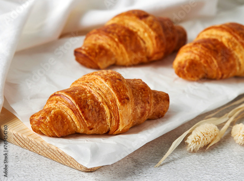 Freshly baked croissants on white baking paper and wooden cutting board with white kitchen towel beside. Close-up shot, homemade food, breakfast concept. Meal serve