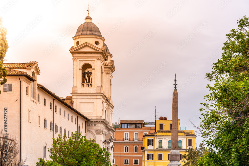 The Trinita dei Monti church stands above the Roman skyline, bathed in the warm glow of a sunset, with an ancient obelisk adding contrast against the pastel-colored buildings.