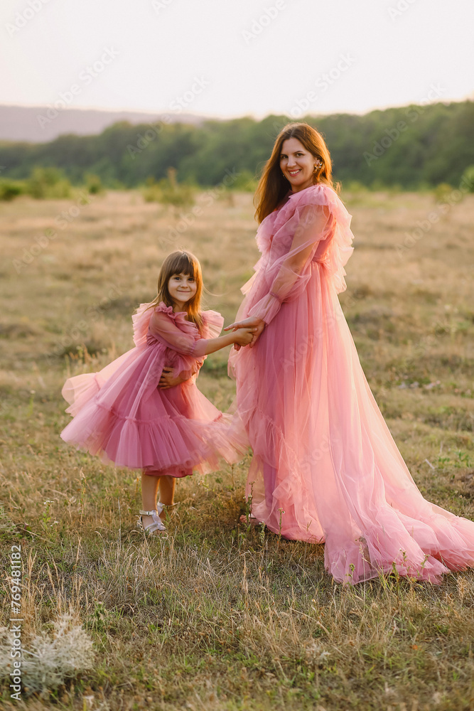 Naklejka premium Cheerful mother and her daughter in tulle pink dresses posing on nature at sunset. Love. Family concept. Photoshooting