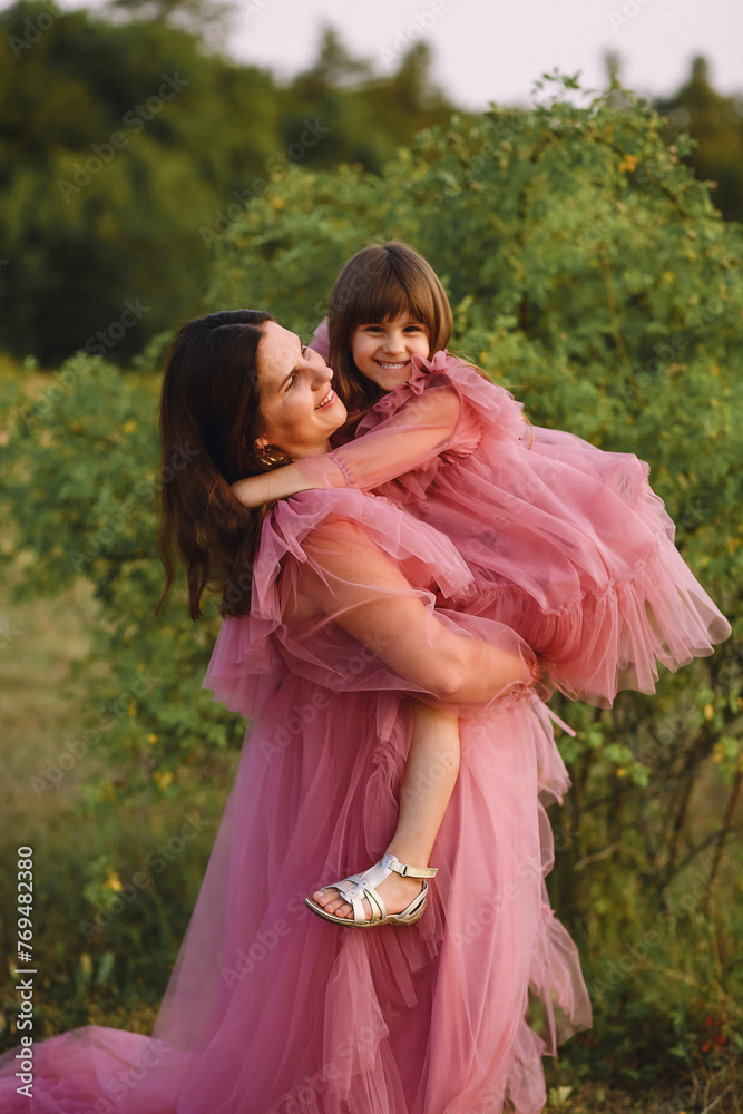 Cheerful mother and her daughter in tulle pink dresses posing outdoor at summer. Beautiful mom holding her little girl in her arms on nature background. Photoshooting Cheerful mother and her daughter in tulle pink dresses posing outdoor at summer. Beautiful mom holding her little girl in her arms on nature background. Photoshooting