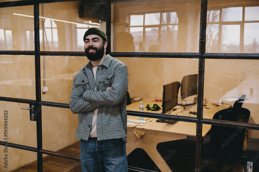 Confident bearded man standing near glass wall while folding arms in office