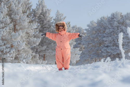 A little girl of 4-6 years old in a pink jumpsuit is smiling at the camera and joyfully raising her hands up, a winter pine forest in the background. Frosty weather