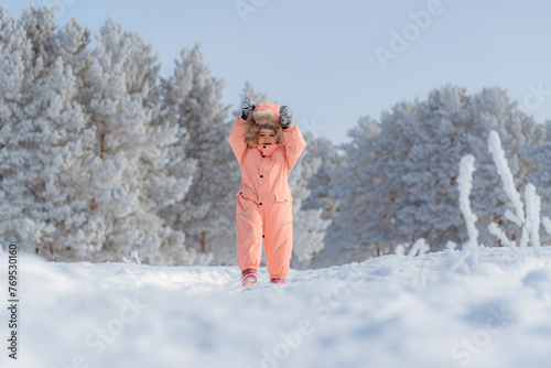 A little girl of 4-6 years old in a pink jumpsuit depicts rabbit ears with her hands to her head, a winter pine forest in the background. Frosty weather