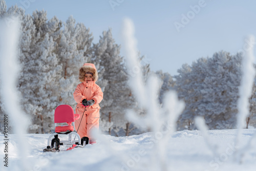 A little 4-6-year-old girl in a pink jumpsuit drags a snowmobile in the middle of a winter park. Copy space