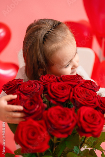 A little girl of 4-6 years old hugs a bouquet of red roses, pink background.